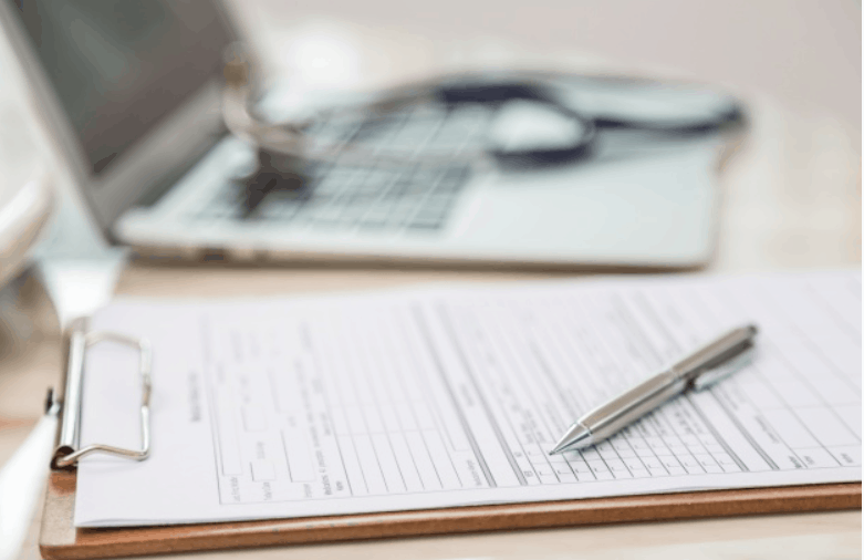 Clipboard with a medical form and pen in front of a laptop and stethoscope
