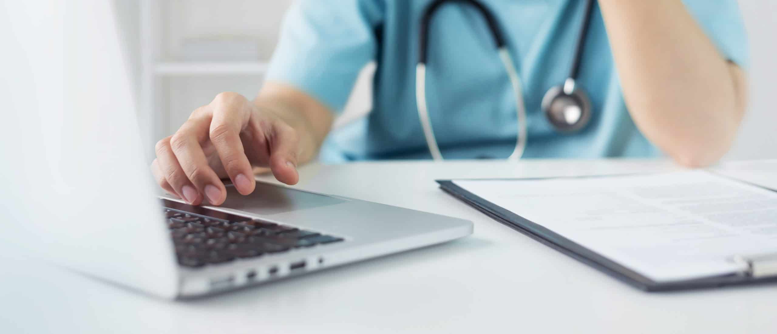 Healthcare worker in scrubs with a stethoscope typing on a laptop beside paperwork
