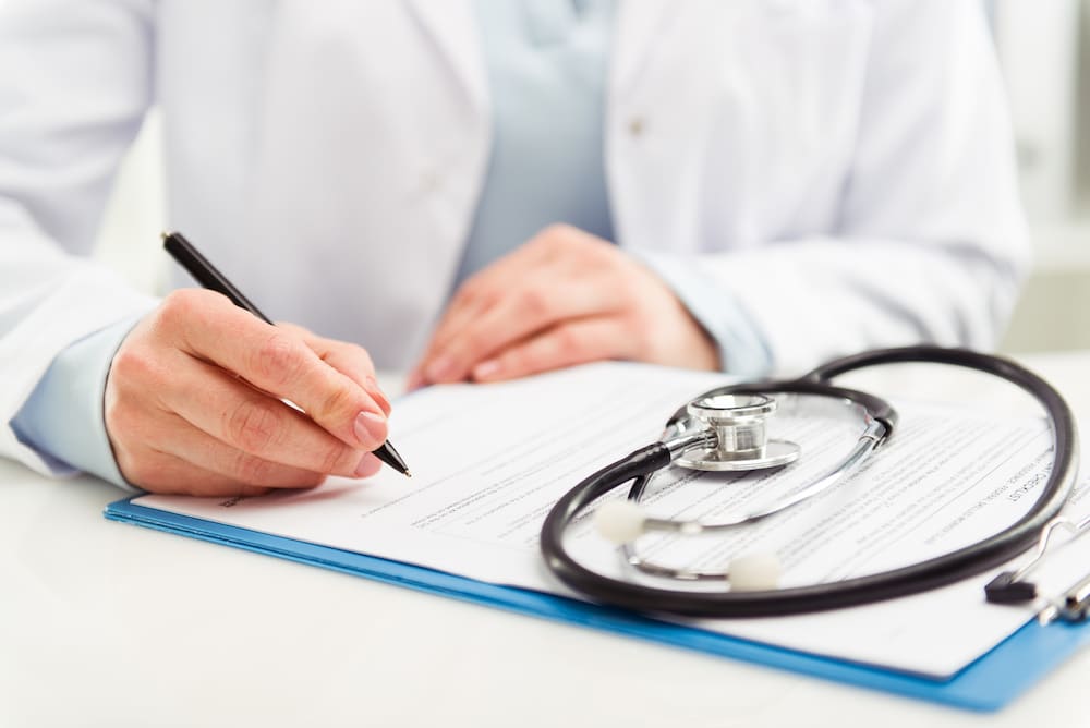 Hand signing a form on a clipboard beside a stethoscope
