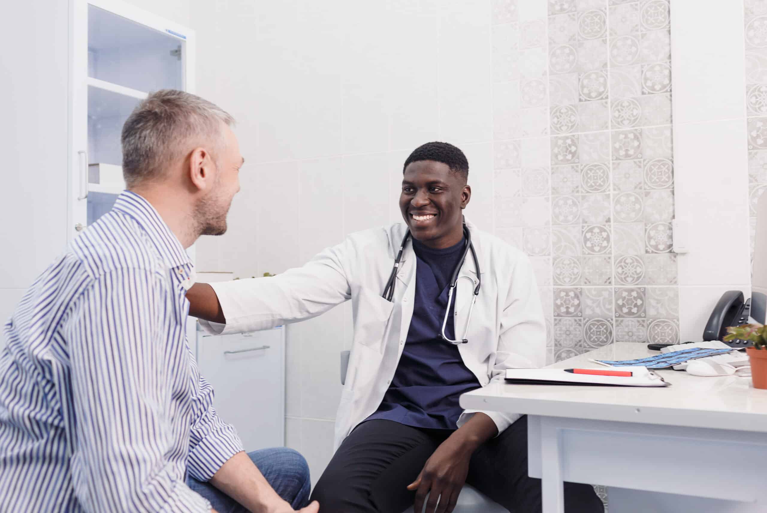 Smiling doctor in a white coat with his hand on a patient's shoulder in an exam room