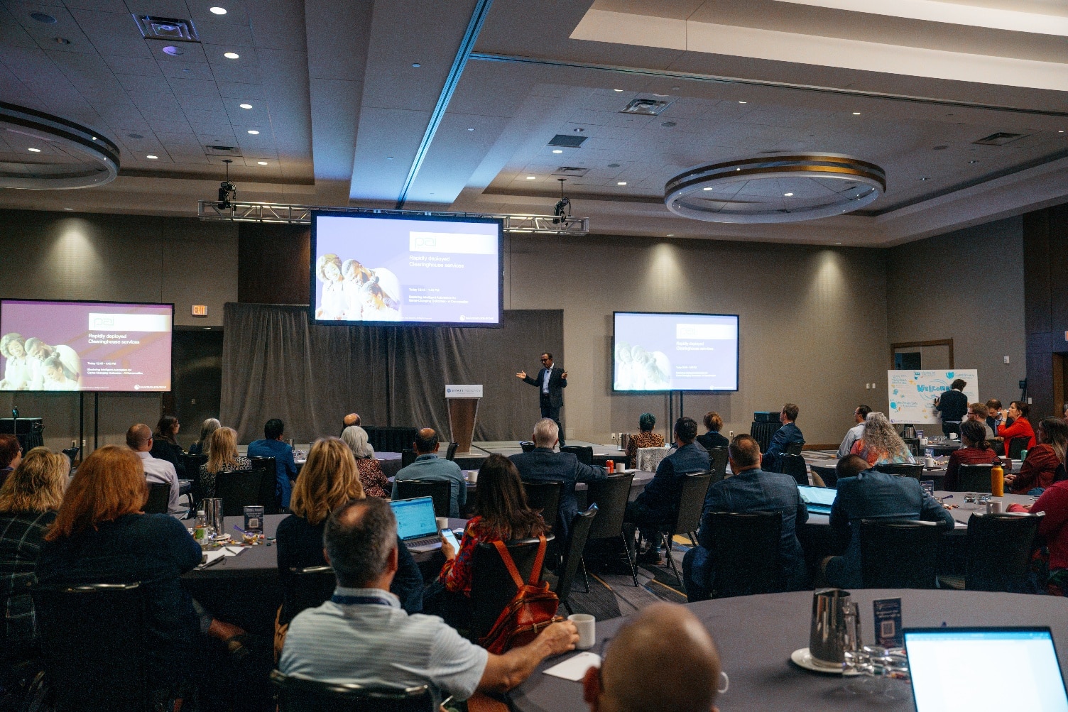 Speaker presenting to seated attendees in a hotel ballroom with screens on stage