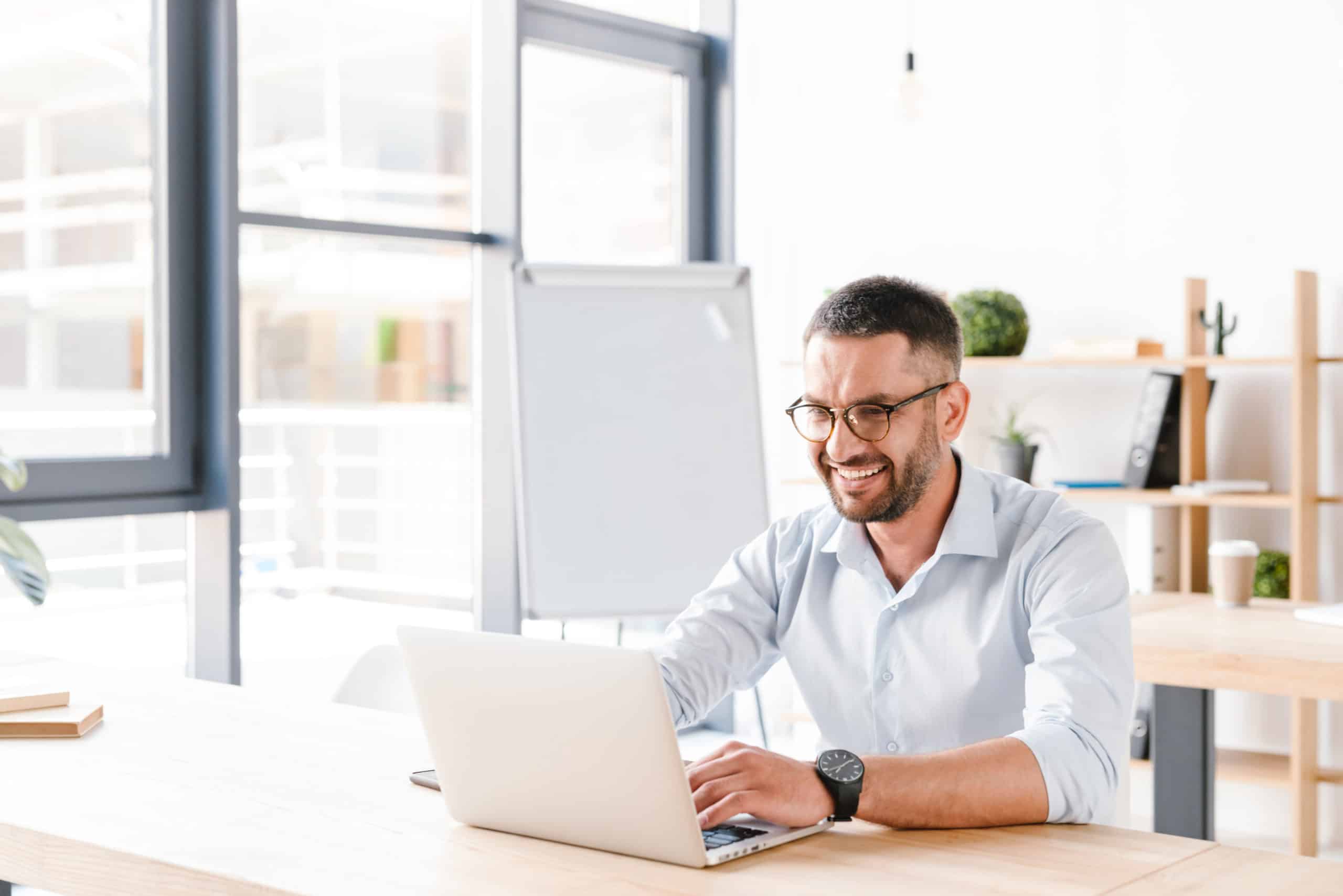 Smiling man with glasses working on a laptop at a desk in a bright office