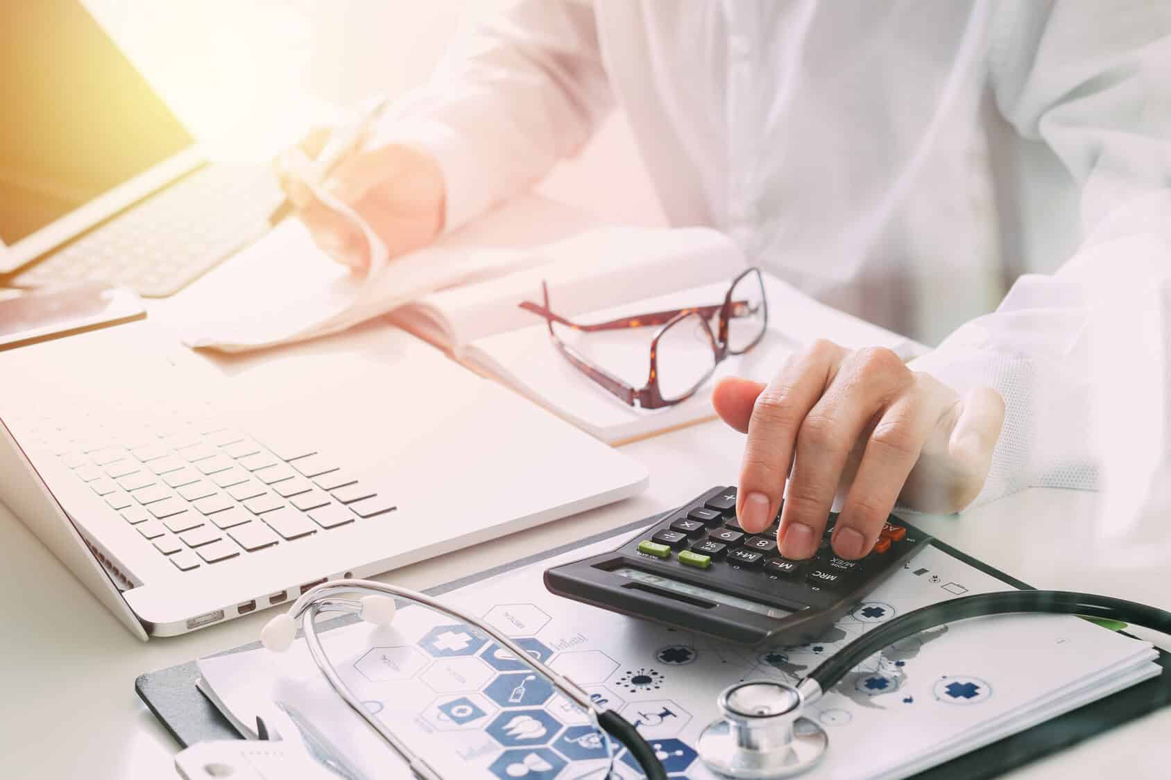 Hand using a calculator at a desk with a laptop, stethoscope and paperwork