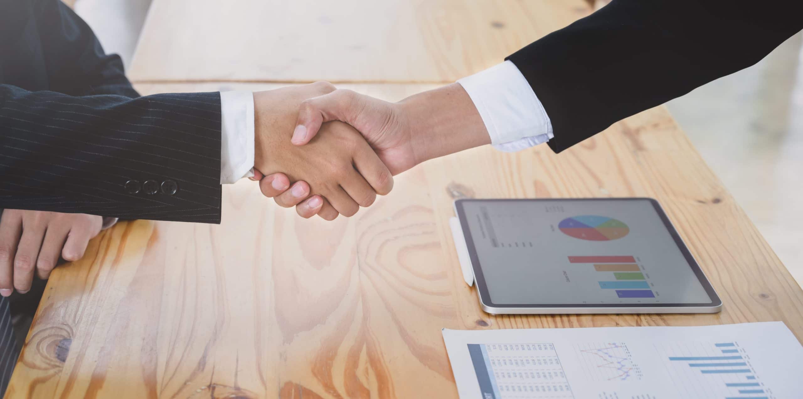 Two business professionals shaking hands across a table with charts on a tablet